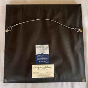 May include: The back of a framed artwork, featuring a dark brown backing. A wire is strung between two metal D-rings for hanging. Two labels are visible: "UltraVue UV70" and "Designers Gallery Fine Art and Custom Framing."