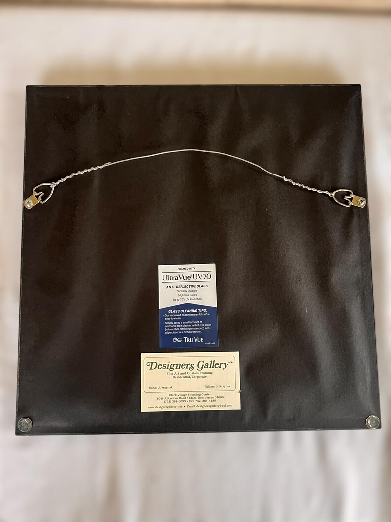 May include: The back of a framed artwork, featuring a dark brown backing. A wire is strung between two metal D-rings for hanging. Two labels are visible: "UltraVue UV70" and "Designers Gallery Fine Art and Custom Framing."