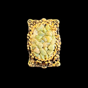 May include: A rectangular gold-toned ring with a central oval of light green stones. The ring has an ornate, textured border and a dark background, highlighting the jewelry's details.