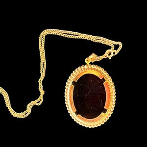 Peut inclure: Collier dor&eacute; avec un pendentif ovale. Le pendentif a un centre sombre et une bordure perl&eacute;e d&eacute;corative. La cha&icirc;ne est de style gourmette. Le collier est pos&eacute; sur un fond noir.