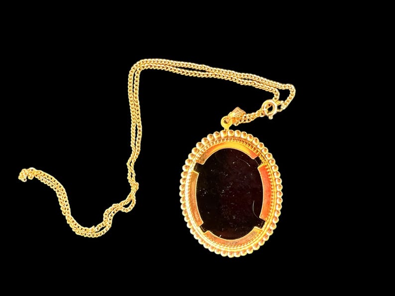 Peut inclure: Collier dor&eacute; avec un pendentif ovale. Le pendentif a un centre sombre et une bordure perl&eacute;e d&eacute;corative. La cha&icirc;ne est de style gourmette. Le collier est pos&eacute; sur un fond noir.