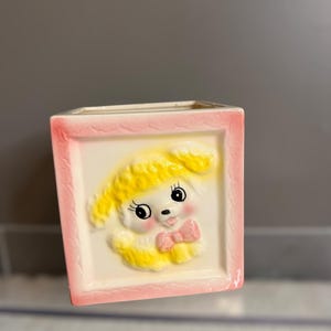 Könnte beinhalten: Eine rechteckige Keramikvase mit rosa Rand und einem erhabenen Relief eines Cartoon-Welpen. Der Welpe hat gelbes Fell, eine rosa Fliege und schwarze Augen. Der Hintergrund ist cremefarben.