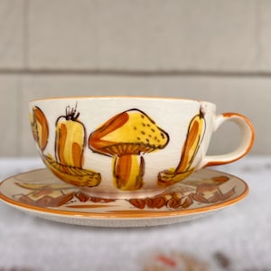 May include: A white ceramic teacup and saucer with a mushroom design. The cup has a handle and is decorated with yellow and brown mushrooms. The saucer has a matching design.