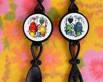 May include: Two black metal kitchen utensils: a spoon and a fork. Each has a decorative handle with a heart design and a round ceramic tile with a colourful teapot and teacup illustration. The background is a floral pattern.