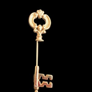 Op de afbeelding: Een goudkleurige sleutelspeld met een sierlijk ontwerp. De speld heeft een sleutelvorm met een decoratieve bovenkant en een lange, slanke speld. De details van de sleutel omvatten een afgeronde bovenkant, een centraal ontwerp en een sleutelgatvorm.