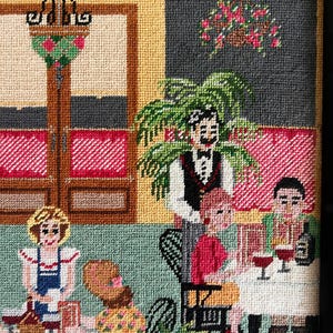 May include: Needlepoint artwork depicting a restaurant scene. The image shows a waiter, patrons, tables, and decorative elements. The color palette includes red, green, yellow, and brown, creating a cozy atmosphere. The artwork is detailed and textured.