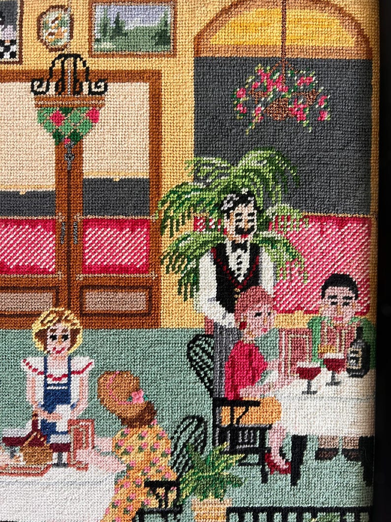 May include: Needlepoint artwork depicting a restaurant scene. The image shows a waiter, patrons, tables, and decorative elements. The color palette includes red, green, yellow, and brown, creating a cozy atmosphere. The artwork is detailed and textured.
