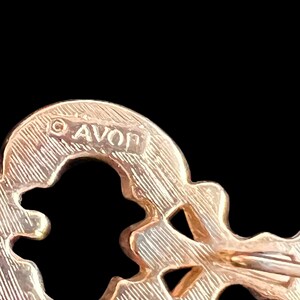 Op de afbeelding: Een goudkleurige sleutelvormige broche met het woord "AVON" bovenaan gestempeld. De broche heeft een getextureerd oppervlak en een sluiting met speld aan de achterkant. Het sleutelontwerp heeft uitgesneden vormen.