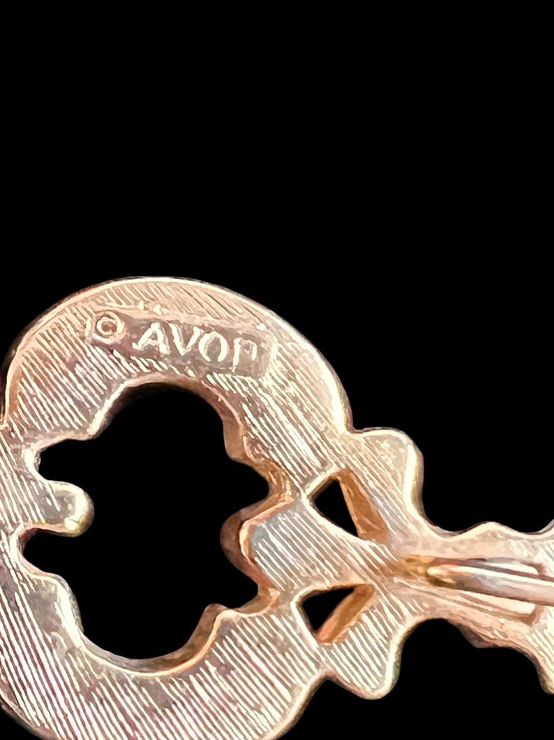 Op de afbeelding: Een goudkleurige sleutelvormige broche met het woord "AVON" bovenaan gestempeld. De broche heeft een getextureerd oppervlak en een sluiting met speld aan de achterkant. Het sleutelontwerp heeft uitgesneden vormen.
