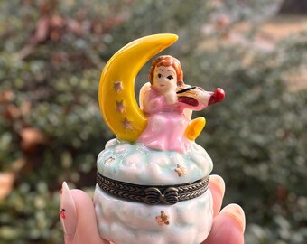 May include: A small, decorative trinket box featuring a celestial theme. The box is shaped like a cloud and topped with a yellow crescent moon and a small angel playing a violin. The angel wears a pink dress. The box is held in a hand.