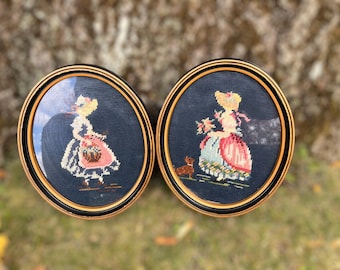 May include: Two framed oval needlepoint artworks. Each features a woman in a dress, with a basket or flowers, and a small dog. The needlepoint is set against a dark blue background, framed in black and gold.
