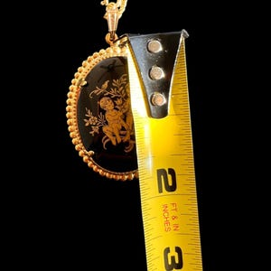Peut inclure: Collier pendentif dor&eacute; avec un centre ovale noir orn&eacute; d'un motif de ch&eacute;rubin dor&eacute;. Le pendentif mesure environ 5 cm de long, avec une bordure perl&eacute;e et une cha&icirc;ne d&eacute;licate.