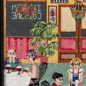 May include: A framed needlepoint scene depicting an outdoor cafe. The artwork features patrons seated at tables, a chalkboard sign with the words "Creative I Cuisine", and floral accents. The color palette includes shades of green, red, and beige.