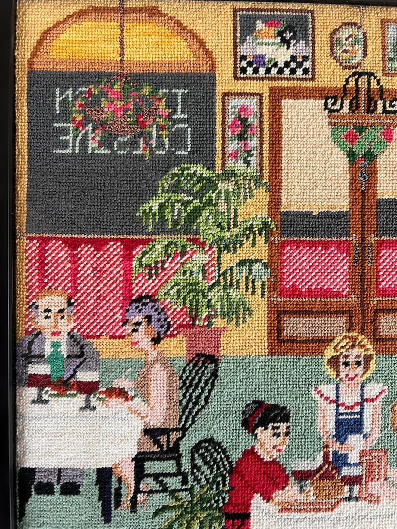May include: A framed needlepoint scene depicting an outdoor cafe. The artwork features patrons seated at tables, a chalkboard sign with the words "Creative I Cuisine", and floral accents. The color palette includes shades of green, red, and beige.