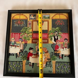 May include: Framed needlepoint art depicting a restaurant scene with patrons and servers. The black frame surrounds the artwork, which measures approximately 12 inches tall. The scene includes tables, chairs, and decorative elements.
