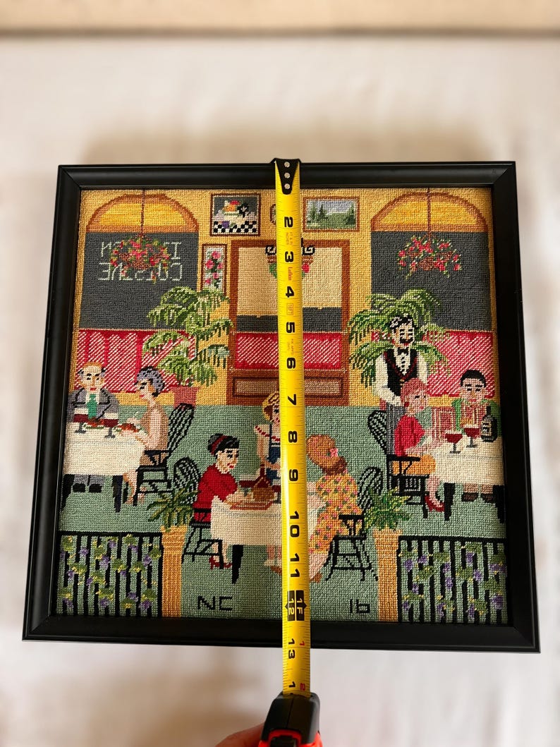 May include: Framed needlepoint art depicting a restaurant scene with patrons and servers. The black frame surrounds the artwork, which measures approximately 12 inches tall. The scene includes tables, chairs, and decorative elements.