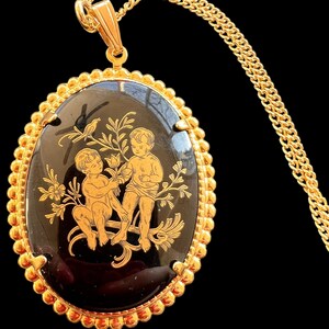 Peut inclure: Collier dor&eacute; avec un pendentif ovale. Le pendentif pr&eacute;sente un fond noir avec une bordure dor&eacute;e et un motif de deux ch&eacute;rubins et d'&eacute;l&eacute;ments floraux. La cha&icirc;ne est &eacute;galement dor&eacute;e, cr&eacute;ant un look classique et &eacute;l&eacute;gant.