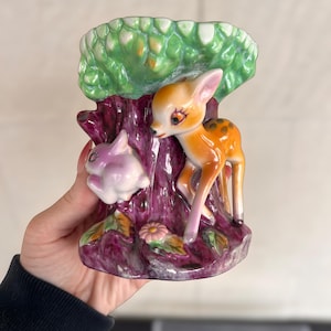 May include: A vintage ceramic vase featuring a fawn and a rabbit near a tree trunk. The vase has a green leafy top, a purple trunk, and a pink and orange fawn. A pink rabbit is on the left side. The base has a flower and leaves.