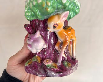 May include: A vintage ceramic vase featuring a fawn and a rabbit near a tree trunk. The vase has a green leafy top, a purple trunk, and a pink and orange fawn. A pink rabbit is on the left side. The base has a flower and leaves.