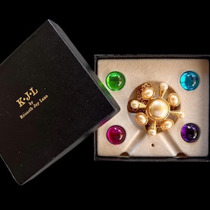 May include: A gold-tone brooch with cream-colored faux pearls in a black box labeled K.J.L by Kenneth Jay Lane. The set includes four interchangeable round cabochon stones in green, blue, magenta, and purple, resting in a beige velvet insert.