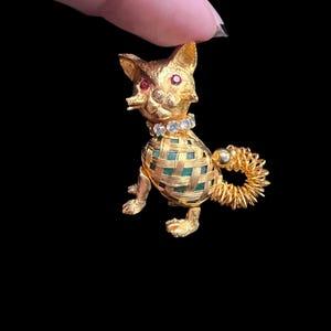 May include: A gold-toned cat brooch featuring a textured metal body with a green woven cage overlay, a collar of clear rhinestones, and two small red rhinestone eyes. The cat has a coiled spring-like tail and a textured, metallic finish.