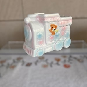 May include: A ceramic train figurine in pastel colors. The train is white with pink and blue accents, featuring a teddy bear in the window. The wheels and decorative clouds are blue. A charming decorative piece.