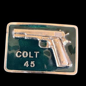 May include: A rectangular belt buckle with a silver-colored pistol design on a dark green background. The text "COLT 45" is displayed in silver lettering. The buckle has a silver border.