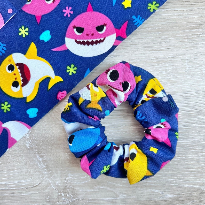 Pu&ograve; includere: Un elastico per capelli blu navy con un motivo ripetitivo di piccoli squali rosa e gialli. L'elastico &egrave; realizzato in tessuto ed &egrave; progettato per tenere i capelli in posizione.