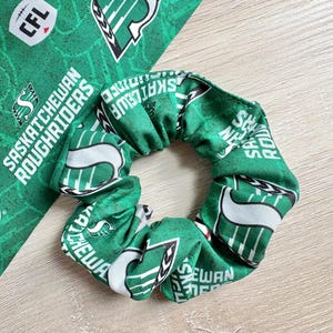 May include: Green scrunchie with white and black accents, featuring the text "Saskatchewan Roughriders" and team logos. A matching fabric piece is visible in the background, displaying the same pattern. The scrunchie is a hair accessory.