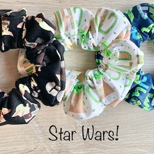 May include: Three Star Wars themed scrunchies. One is black with a white and green pattern of Baby Yoda characters. One is white with a brown and green pattern of Baby Yoda characters. One is blue with a white and green pattern of Baby Yoda characters. The text "Star Wars!" is written below the scrunchies.