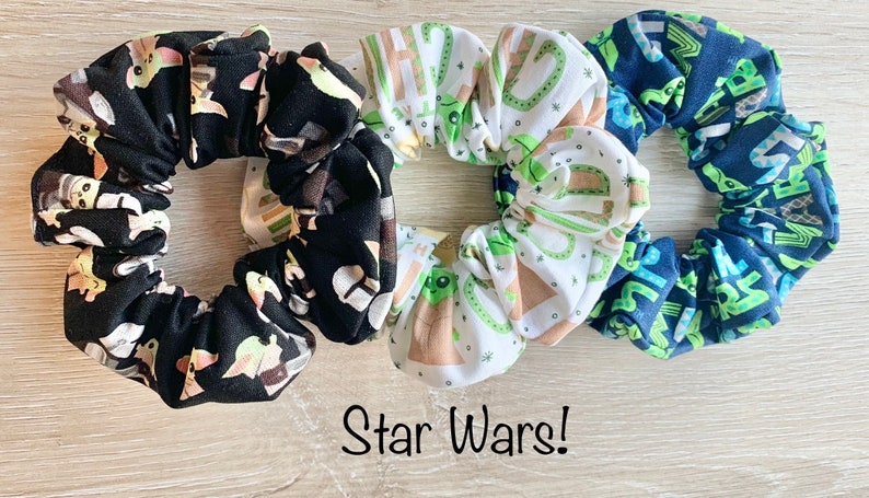 May include: Three Star Wars themed scrunchies. One is black with a white and green pattern of Baby Yoda characters. One is white with a brown and green pattern of Baby Yoda characters. One is blue with a white and green pattern of Baby Yoda characters. The text "Star Wars!" is written below the scrunchies.