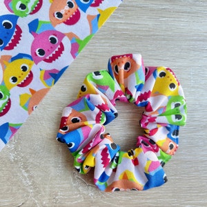 Pu&ograve; includere: Un elastico per capelli bianco con un motivo colorato di personaggi di baby shark. L'elastico &egrave; fatto di tessuto e ha un aspetto raccolto.