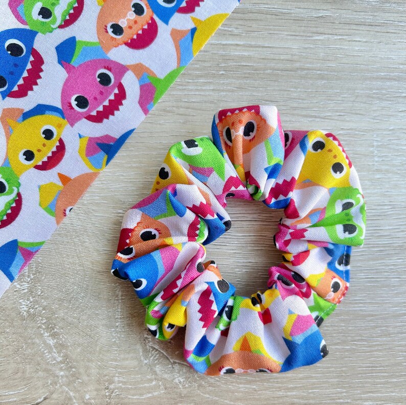 Pu&ograve; includere: Un elastico per capelli bianco con un motivo colorato di personaggi di baby shark. L'elastico &egrave; fatto di tessuto e ha un aspetto raccolto.