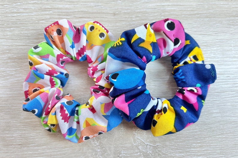 Pu&ograve; includere: Due scrunchies per capelli colorati con un motivo di baby shark. Uno scrunchie &egrave; bianco con baby shark rosa, blu, giallo e verde. L'altro scrunchie &egrave; blu navy con baby shark rosa, giallo e blu.