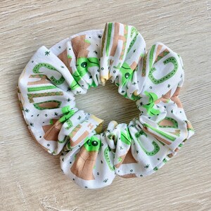 May include: White scrunchie with a green and brown print of a cartoon character. The character is surrounded by stars.
