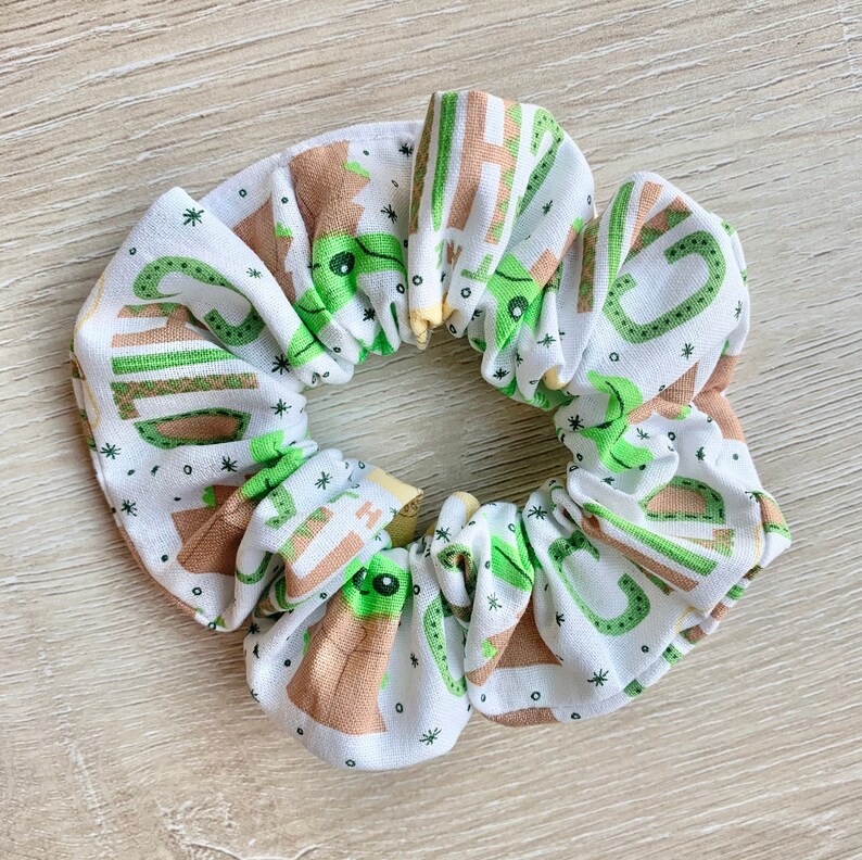 May include: White scrunchie with a green and brown print of a cartoon character. The character is surrounded by stars.