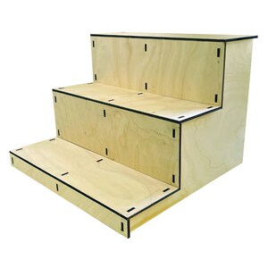 May include: A three-tiered wooden display stand with a light wood grain finish. The edges are outlined with black trim. The stand is designed with three steps, perfect for showcasing products or items. The overall dimensions are approximately 24 inches wide, 18 inches deep, and 12 inches high.