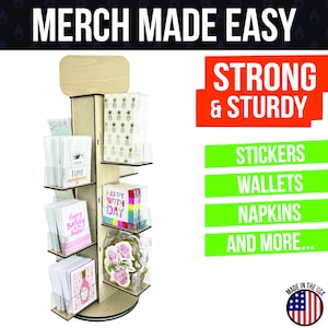 May include: A wooden rotating display stand with six shelves for showcasing merchandise. The shelves are filled with various items, including greeting cards, napkins, and stickers. The stand is labeled "MERCH MADE EASY" and "STRONG & STURDY".