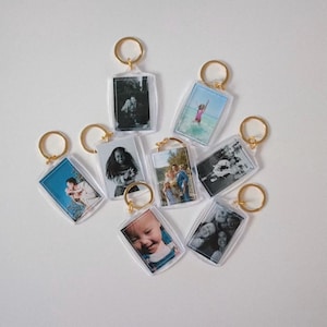 May include: A collection of clear acrylic keychains with gold-colored key rings. Each keychain features a rectangular photo, showcasing various black and white and color images. The keychains are arranged on a white surface.