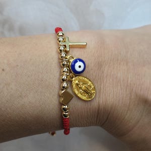 Puede incluir: Una pulsera de hilo rojo con cuentas y dijes dorados. Los dijes incluyen una cruz, un ojo azul, un coraz&oacute;n y un medall&oacute;n dorado. La pulsera se lleva en la mu&ntilde;eca.