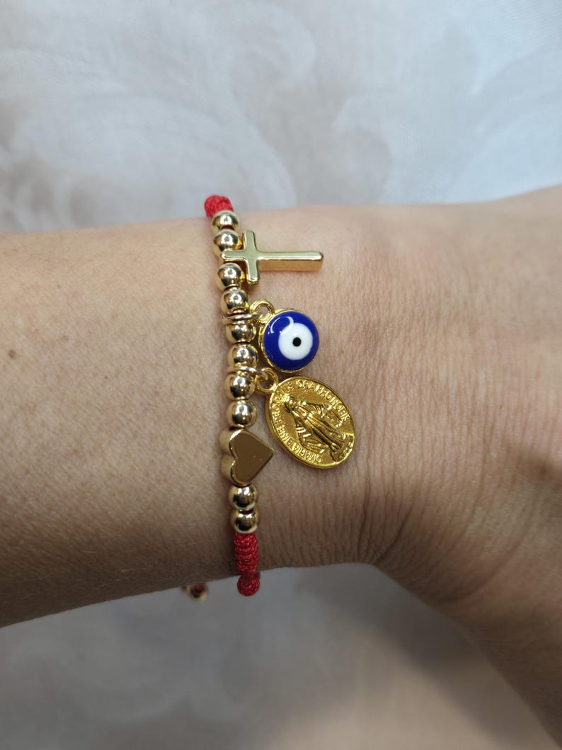 Puede incluir: Una pulsera de hilo rojo con cuentas y dijes dorados. Los dijes incluyen una cruz, un ojo azul, un coraz&oacute;n y un medall&oacute;n dorado. La pulsera se lleva en la mu&ntilde;eca.