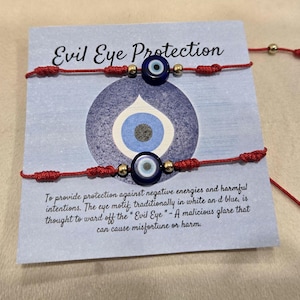 May include: Two red string bracelets with blue and white evil eye beads and gold-colored accents. The bracelets are displayed on a card with the text "Evil Eye Protection" and a description of the evil eye's purpose.