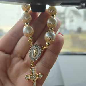 May include: A gold rosary with faux pearl beads and a cross pendant. The cross is encrusted with clear crystals. The rosary features a gold oval pendant with a religious figure. The beads are white and gold.