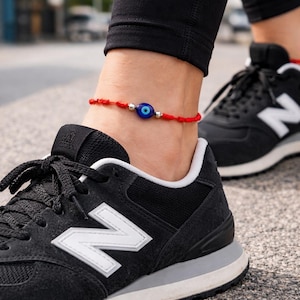 May include: Red string ankle bracelet with a blue and white evil eye charm. The bracelet is worn on an ankle, paired with black sneakers with white accents. The shoes are on a paved surface.