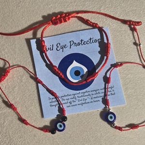 May include: Red string bracelets with adjustable knots and small blue evil eye charms. A card with the text "Evil Eye Protection" is also present. The bracelets are designed to be worn on the wrist.