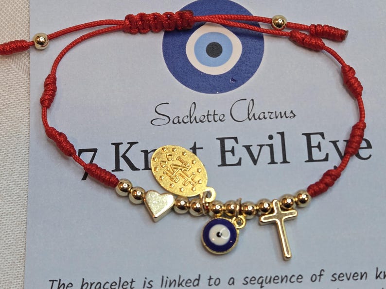 Puede incluir: Pulsera de hilo rojo con cuentas y dijes dorados, incluyendo un coraz&oacute;n, una cruz y un ojo turco. La pulsera tiene un cierre de nudo corredizo. El texto "Sachette Charms" y "7 Knot Evil Eye" son visibles.