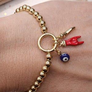 May include: Gold beaded bracelet with a large gold ring clasp and three charms. Charms include a red hand, a gold horn, and a blue evil eye. The bracelet is a stylish accessory.