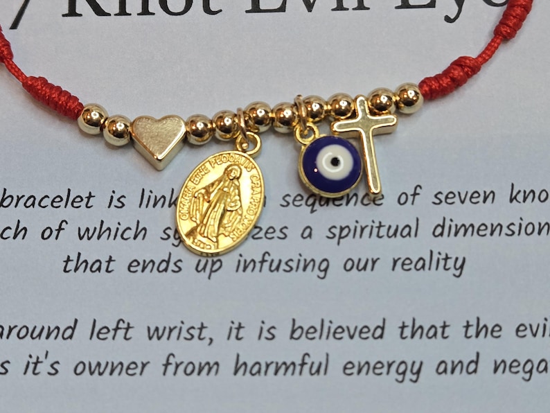 Puede incluir: Pulsera de cord&oacute;n rojo con dijes dorados: un coraz&oacute;n, una medalla religiosa, una cruz y un ojo turco. La pulsera est&aacute; vinculada a una secuencia de siete nudos.