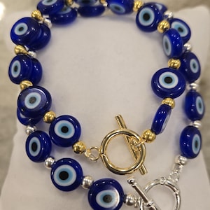 May include: Two bracelets featuring dark blue, eye-shaped beads with white and black details. The bracelets are strung with silver and gold-colored beads and have a gold and silver toggle clasp. The bracelets are displayed on a white surface.