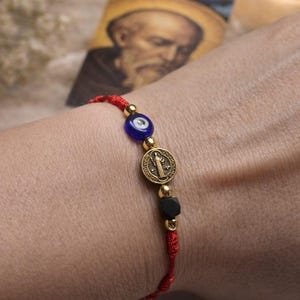 May include: Red cord bracelet featuring gold beads, a blue eye bead, a round gold charm with a religious figure, and a black bead. The bracelet is displayed on a wrist, with a religious image in the background.
